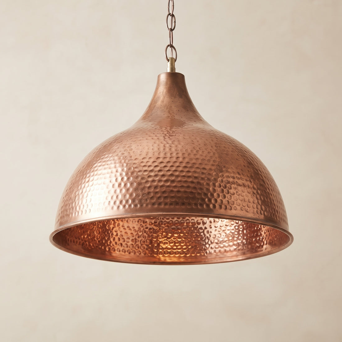A hammered copper pendant light with a dome-shaped shade hanging from a metal chain against a warm beige background.