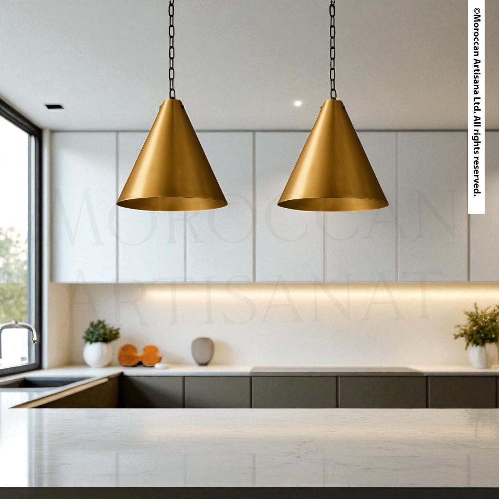 a modern kitchen with a sleek, minimalist design. The focal point is a pair of golden pendant lights hanging above a white countertop, which is set against a backdrop of white cabinets. The kitchen also includes a sink and a few decorative elements, such as potted plants and a vase.